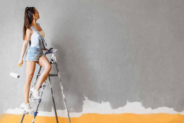 Happy woman paints wall on ladder with roller at home. Repair concept - Fotó, kép