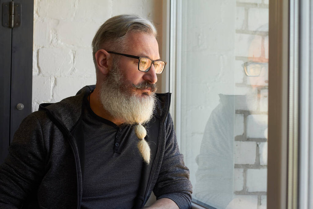 Half-length portrait of mature gray-haired man with original beard on window backdrop, selective focus - Photo, image