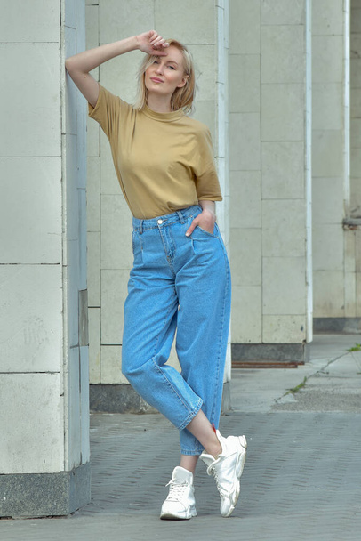 Молодая привлекательная красивая модная стройная женщина стоит рядом с серой стеной в ярко-синих джинсах и бежевой футболке. ветер в волосах. современная женщина в кроссовках. уличный стиль
 - Фото, изображение