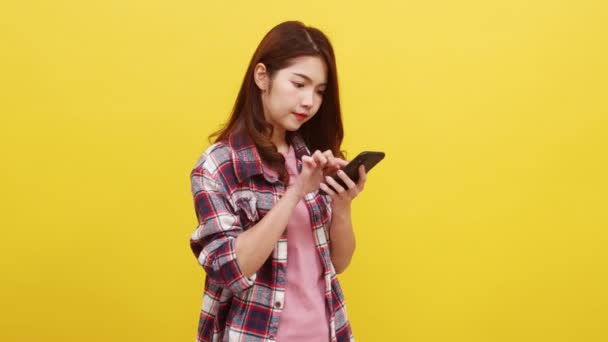Muotokuva yllättynyt aasialainen nainen käyttää matkapuhelinta positiivinen ilme, pukeutunut rento vaatteet ja katsot kameraa keltaisella taustalla. Onnellinen ihana iloinen nainen iloitsee menestyksestä. - Materiaali, video