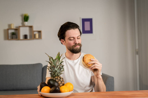 A young man sits at a table and talks about the usefulness of fruits, he picks them up and shows pineapple, avocado, orange. Healthy food - 写真・画像