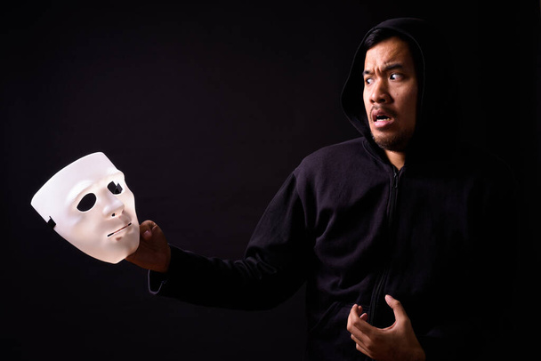 Studio shot of young Asian man with hoodie and mask against black background - 写真・画像