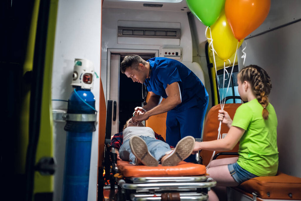 Madre inconsciente acostada en una camilla en un carro de ambulancia, un paramédico brindando sus primeros auxilios y hablando con su pequeño hijo.
 - Foto, imagen