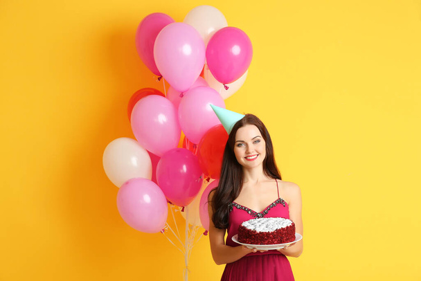 Beautiful young woman in party cap with tasty cake and air balloons on yellow background - Photo, Image