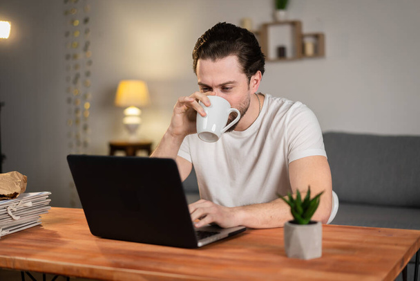 Un giovane lavora da casa e beve tè alla scrivania con il computer. Pessima idea. Liquido in tazza, caffè - Foto, immagini
