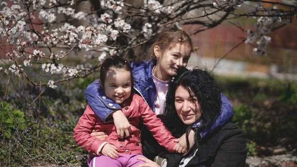 Mom with two cute daughters in a park near a flowering tree in spring. Concept of happy family and spting - Photo, Image