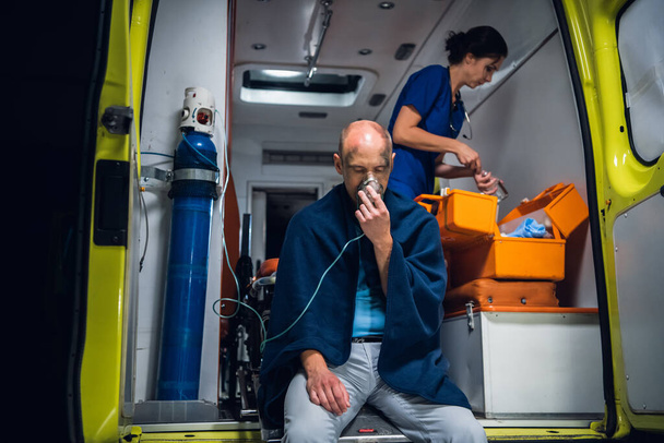 Поранений чоловік у ковдрі, що дихає через кисневу маску в машині швидкої допомоги, медсестра упаковує медичне обладнання на задньому плані
 - Фото, зображення