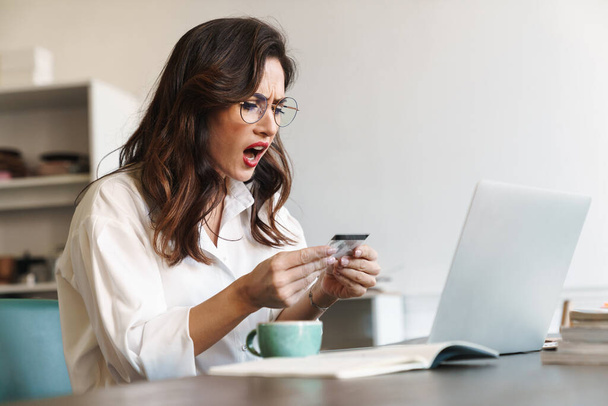 Atraktivní šokovaný naštvaný mladý brunetka businesswoman sedí u jídelního stolu s notebookem uvnitř, nakupování online s kreditní kartou - Fotografie, Obrázek
