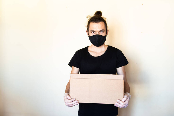 A man wear protective face mask and gloves holding cardboard box. Delivery service and volunteering during pandemic coronavirus concept. - Photo, image