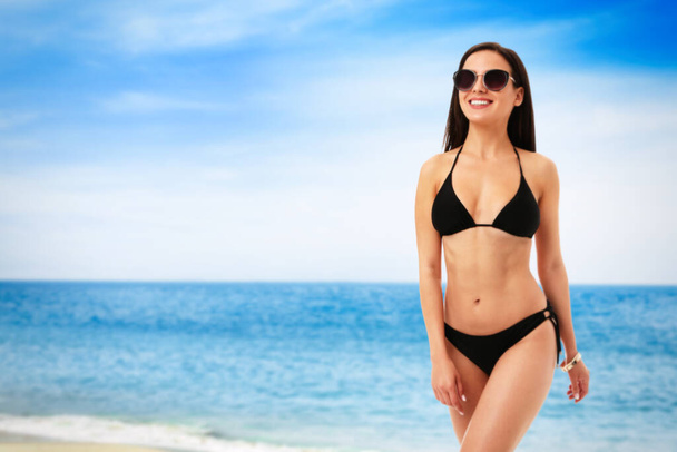 Pretty sexy woman with slim body in stylish black bikini near ocean on sunny day, space for text - Zdjęcie, obraz