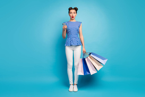 Full length photo of pretty open mouth lady carry packs addicted shopper reading good news telephone wear dotted blouse white pants sneakers isolated blue color background - Фото, изображение