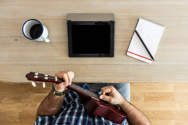 nerozpoznatelný muž nacvičuje doma ukulele s tabletem, notebookem a kávou, pohled shora - Fotografie, Obrázek