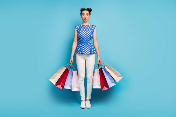 Teljes hosszúságú fotó szép nyitott szájú hölgy hordoznak sok csomag ámokfutás függő vásárló shopaholic kopás pontozott blúz fehér nadrág lábbeli elszigetelt kék háttér - Fotó, kép