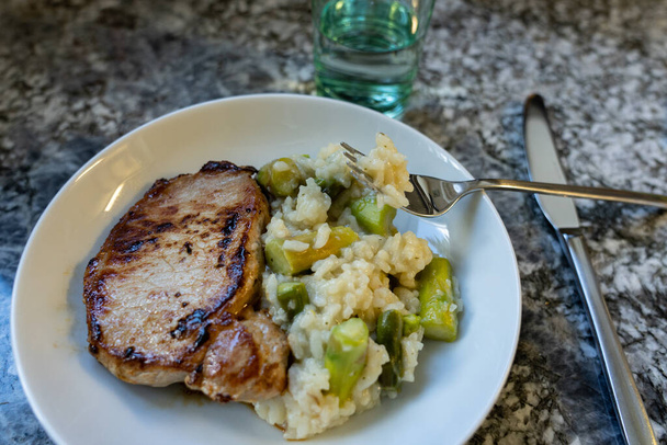 Carne asada y arroz risotto con espárragos en plato redondo blanco con tenedor y cuchillo sobre fondo de granito. Vista de ángulo alto
. - Foto, Imagen