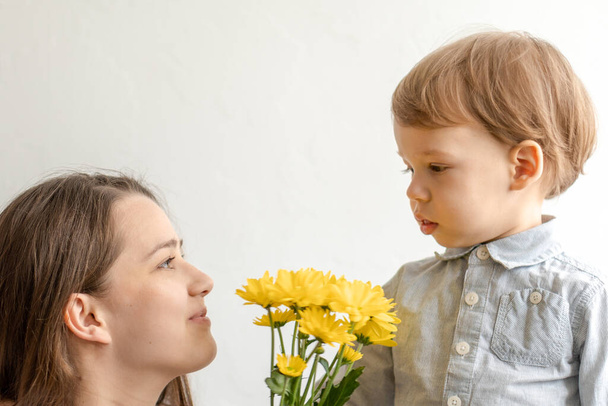 motherhood, mothers day, birthday, childhood, family concept - close-up little boy in blue colorful dress congratulate and give young mother bright bouquet of yellow daisy flowers.on white background. - Фото, изображение