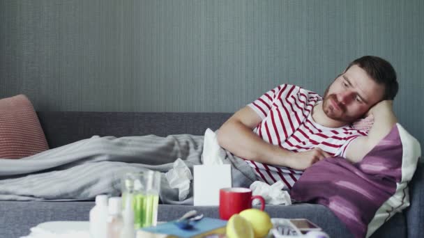 Nemocný muž s chřipkou leží doma na gauči. Nemocný mladý muž se studenou kontrolní teplotou s teploměrem - Záběry, video