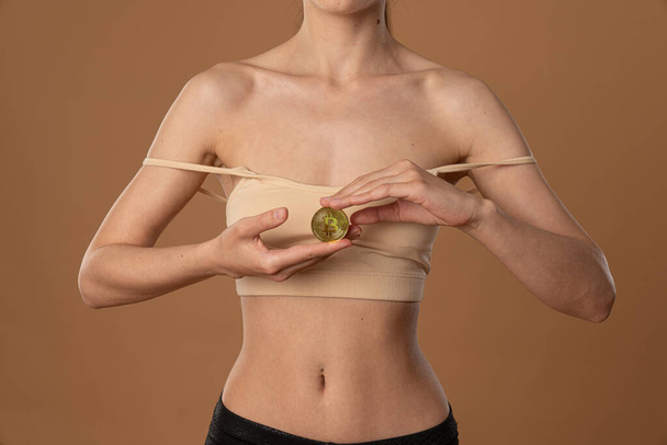Cropped image of a young female in beige bra, with naked shoulders who holds golden bitcoin coin in her hands - Photo, Image