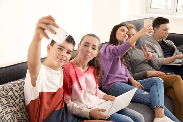 Groupe d'adolescents avec des appareils modernes prenant selfie tout en étant assis sur le canapé à l'intérieur
 - Photo, image