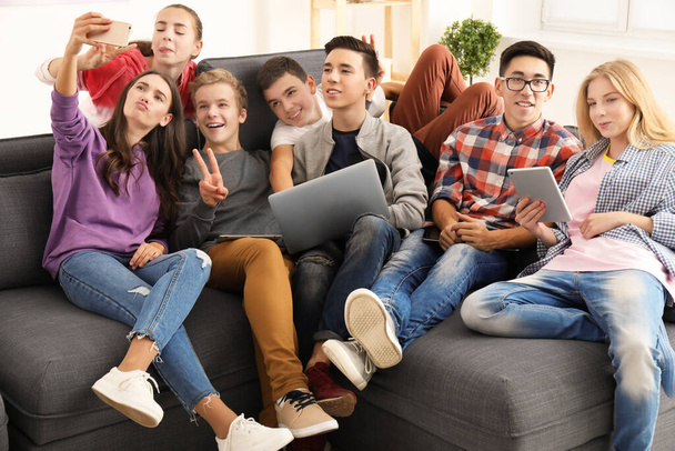 Grupo de adolescentes com dispositivos modernos tomando selfie enquanto se senta no sofá dentro de casa
 - Foto, Imagem