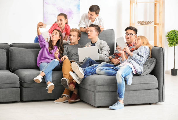 Group of teenagers with modern devices taking selfie while sitting on sofa indoors - Fotografie, Obrázek
