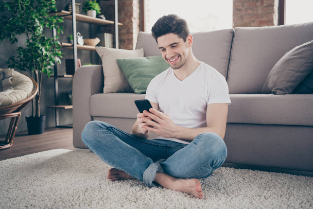 Portrait of his he nice attractive focused glad cheerful cheery guy sitting on carpet using cell browsing media multimedia at modern industrial loft brick interior style living-room - Fotó, kép