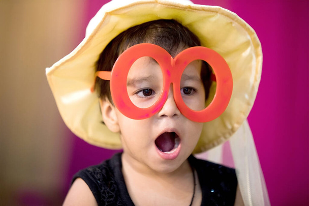 Miúdo indiano vestindo óculos grandes e boné dando expressão Wow com boca aberta durante a festa
 - Foto, Imagem