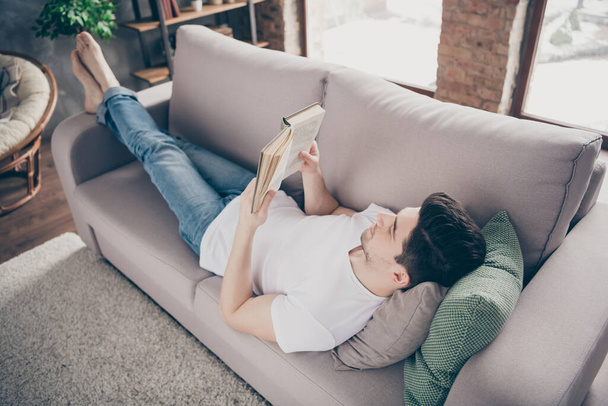 Profile side view portrait of his he nice attractive peaceful focused brunette guy lying on divan reading book spending day at modern industrial loft brick interior style living-room - Photo, image