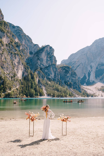 Piękna panna młoda w białej sukience z rękawami i koronką, z żółtym jesiennym bukietem na tle łuku na ceremonię, w Lago di Braies we Włoszech. Ślub przeznaczenia w Europie, nad jeziorem Braies. - Zdjęcie, obraz