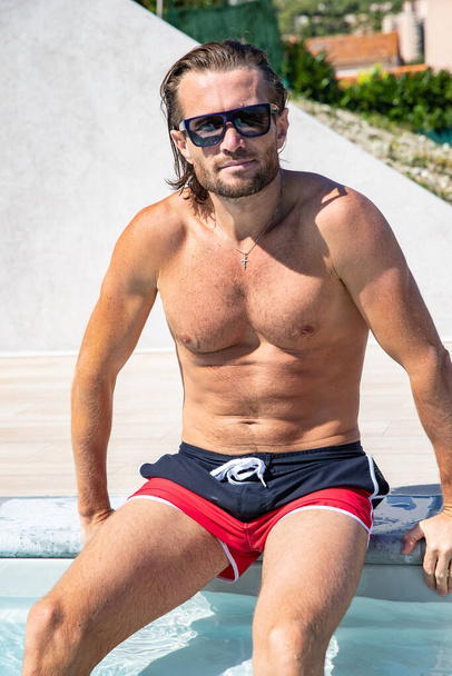 The handsome brutal man in sunglasses with a long hair and naked torso sits near the pool, is looking a camera, a sports suntanned body, sunglasses with a blue frame, he is red blue swimming shorts - Фото, изображение