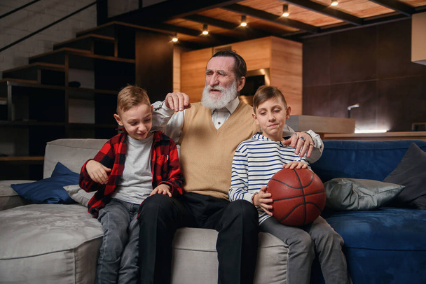 Concentrated senior bearded grandpa with his happy grandsons spending their leisure at revision the basketball championship on tv at home. - Foto, Imagen