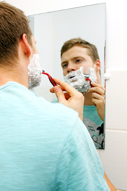 Молодой человек бреет лицо, отражается в зеркале
 - Фото, изображение