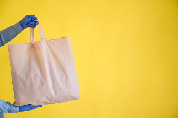 Sending eco-friendly reusable cotton bag on a yellow background. The courier holds a canvas fabric bag without plastic packaging on a yellow background. Safe delivery to the epidemic. - Photo, Image