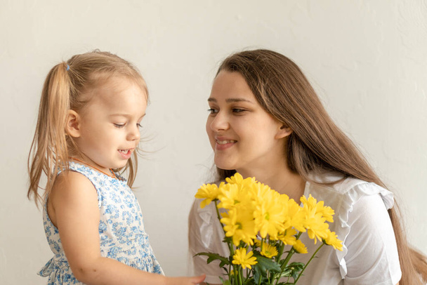 motherhood, mothers day, birthday, childhood, family concept - close-up little girl in blue colorful dress congratulate and give young mother bright bouquet of yellow daisy flowers.on white background - Фото, изображение