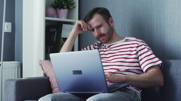 Tired Young Man on a Laptop Holding it on His Lap and Sitting on a Sofa at Home. - Imágenes, Vídeo