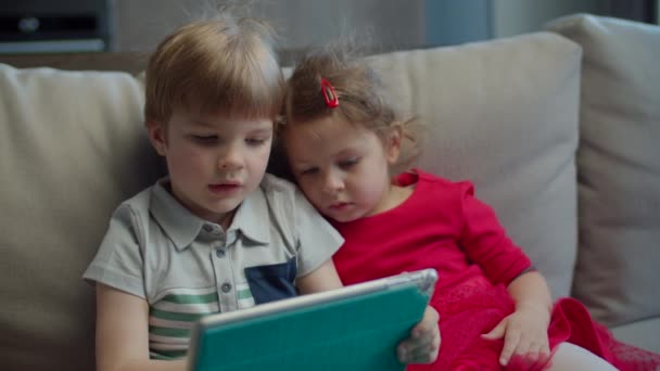 Preschool boy and girl using tablet computer sitting on couch at home. Kids using mobile application on tablet computer together.  - Metraje, vídeo