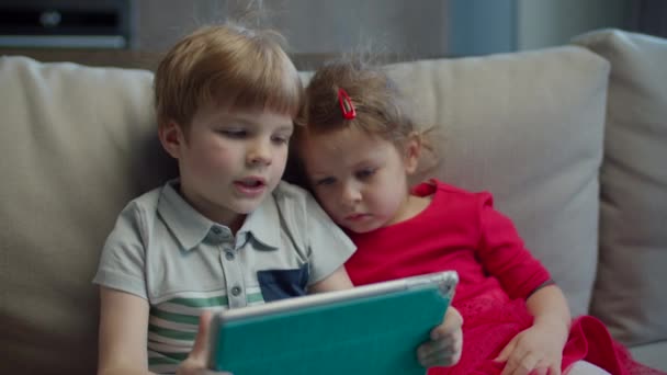 Preschool boy and girl using tablet computer sitting on couch at home. Kids using mobile application on tablet computer together.  - Кадри, відео