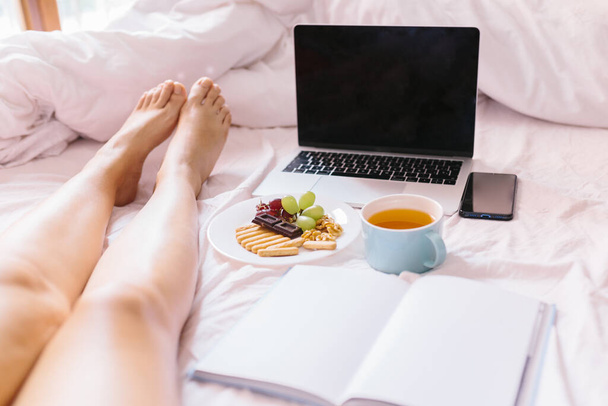 Nohy mladé ženy. Čaj doma v posteli, kontrola notebooku, čtení knihy a snídaní. - Fotografie, Obrázek