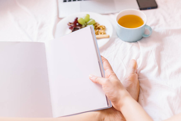 若い女性が本を読んでいる。彼女のベッドで家でお茶,彼女のラップトップをチェック,本を読んで朝食を持っています. - 写真・画像
