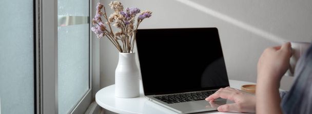 Cropped shot of female freelancer working on laptop with flower vase on white  circle coffee table at home - Fotó, kép