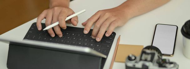 Fotografía recortada del fotógrafo escribiendo en la tableta digital mientras está sentado en la mesa de trabajo con bloc de notas y cámara
 - Foto, Imagen