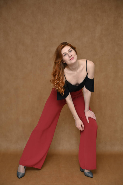 Model posing with long beautiful red hair. Photo full-length portrait on a beige background Caucasian pretty young woman in a black blouse, red trousers. Universal concept. - Foto, immagini