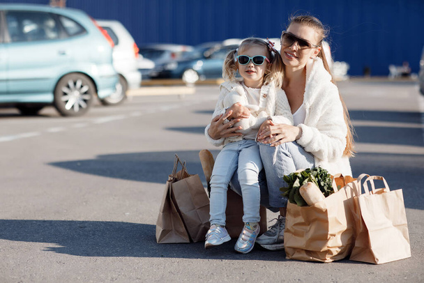Mom and little daughter with ponytails, dressed in white T-shirts and blue jeans posing in the background of the parking lot near the supermarket with packages in hand. Mother and daughter shopping outdoors. - Zdjęcie, obraz
