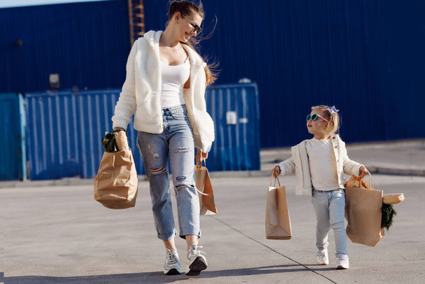 Mutter und kleine Tochter mit Pferdeschwanz, gekleidet in weiße T-Shirts und blaue Jeans posieren mit Paketen in der Hand im Hintergrund auf dem Parkplatz neben dem Supermarkt. Mutter und Tochter beim Einkaufen im Freien. - Foto, Bild