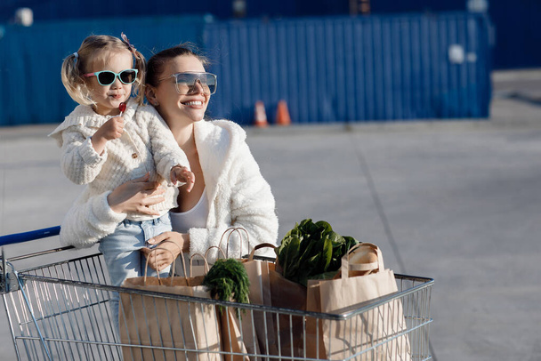 Mom and little daughter with ponytails, dressed in white T-shirts and blue jeans posing in the background of the parking lot near the supermarket with packages in hand. Mother and daughter shopping outdoors. - Foto, imagen