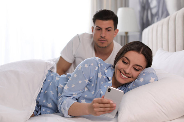Distrustful man peering into girlfriend's smartphone at home. Jealousy in relationship - Fotografie, Obrázek