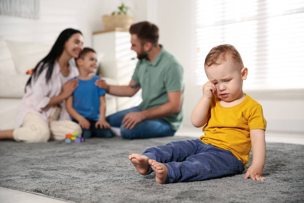 Unhappy baby sitting alone on floor while parents spending time with his elder brother at home. Jealousy in family - Фото, изображение