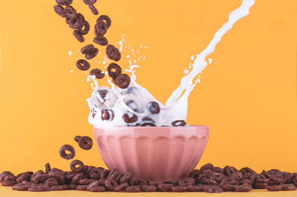 黄色の背景にミルクカップ,チョコレートシリアルが周りに落ち、ミルクが飛び散ると. - 写真・画像