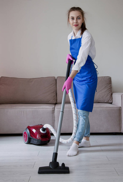 A beautiful young girl in blue jeans in full growth is vacuuming a room near a beige sofa. vertical photo. - Foto, immagini