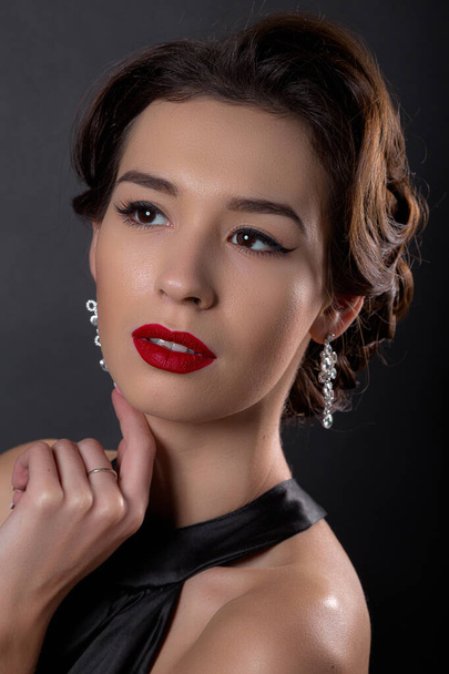 Beautiful gorgeous woman with elegant wavy hairstyle, bright make up. Fashion brunette wearing black dress posing isolated on black background. Classic makeup in eyelid arrows and red lipstick - Photo, image