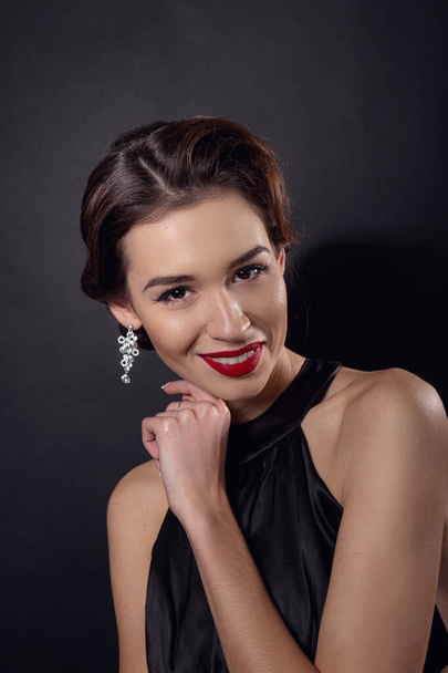 Beautiful gorgeous woman with elegant wavy hairstyle, bright make up. Fashion brunette wearing black dress posing isolated on black background. Classic makeup in eyelid arrows and red lipstick - Fotoğraf, Görsel